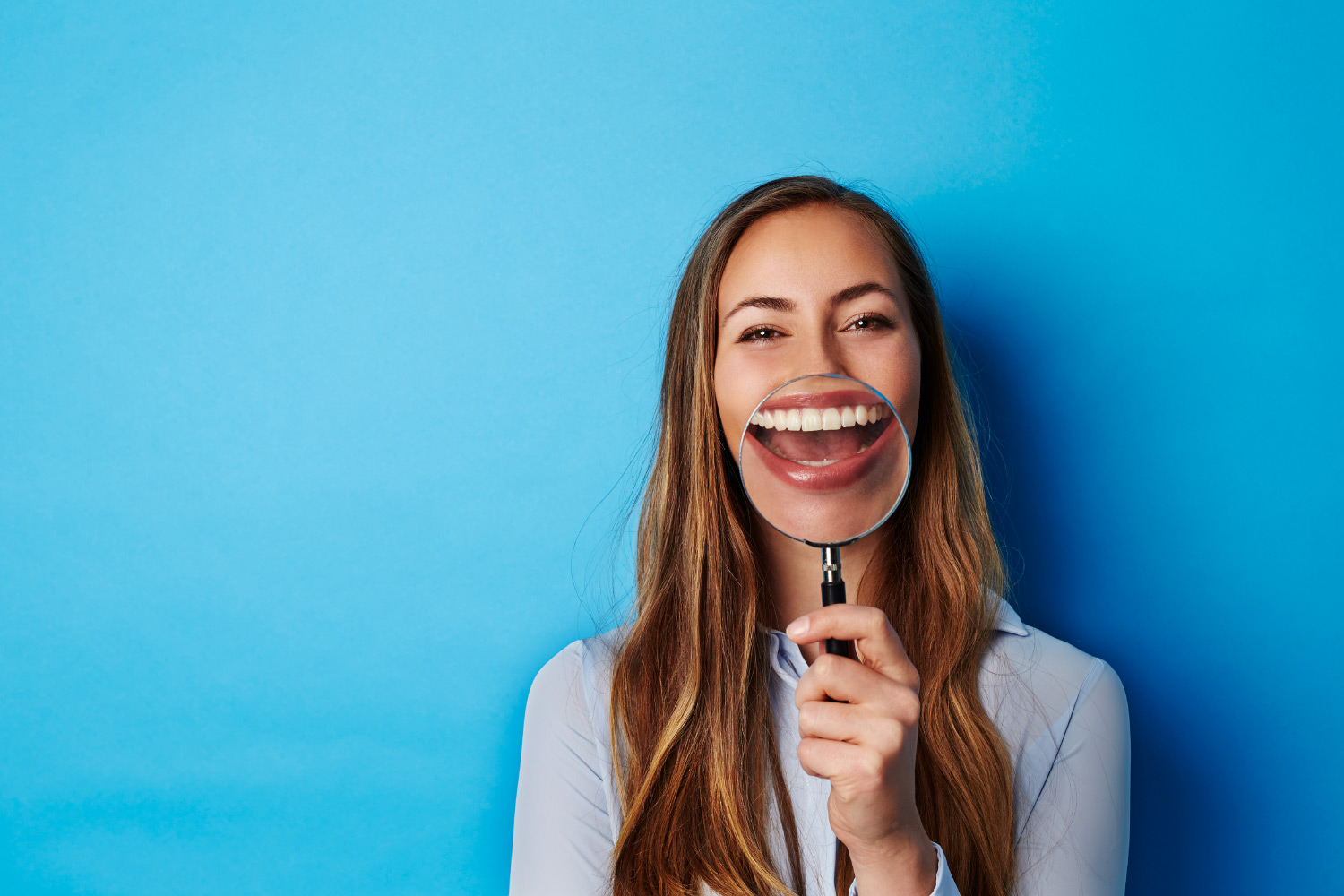 A brunette woman smiles as she holds a magnifying glass to her mouth against a blue wall
